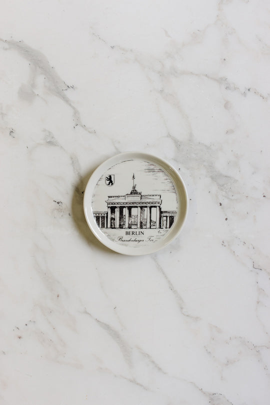 A german coaster, small and round, features a black-and-white illustration of Berlin’s Brandenburg Gate with "Berlin" written below, displayed on a white marble surface.