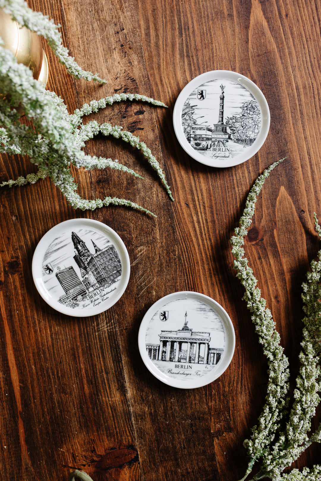 Three small round german coasters featuring black-and-white sketches of Berlin landmarks rest on a wooden surface, surrounded by white flowers and greenery, emphasizing their compact size and decorative charm.