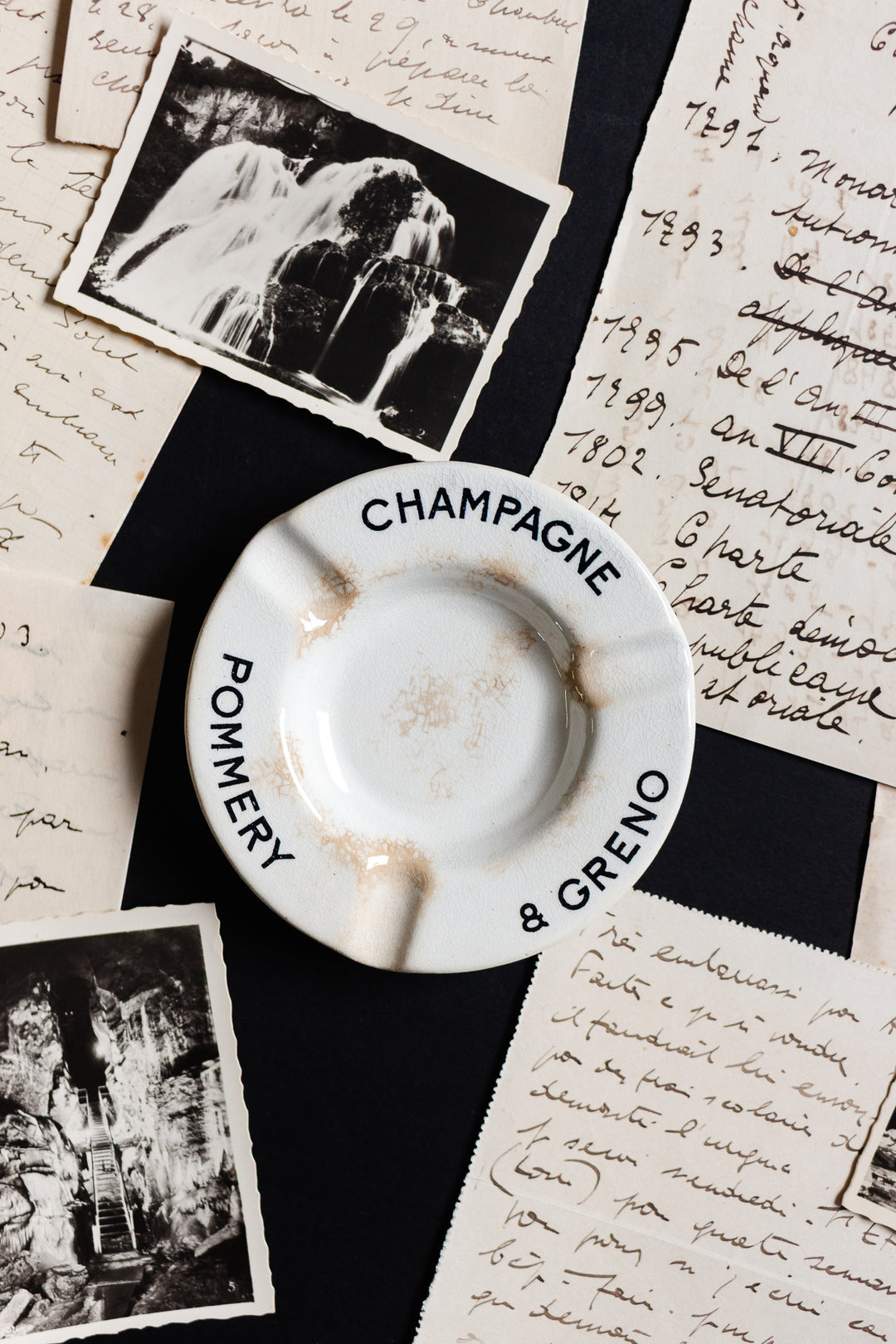 An antique French Luneville Champagne Pommery advertising ashtray rests on handwritten letters and vintage photos, evoking a nostalgic, timeless elegance and a sense of history.