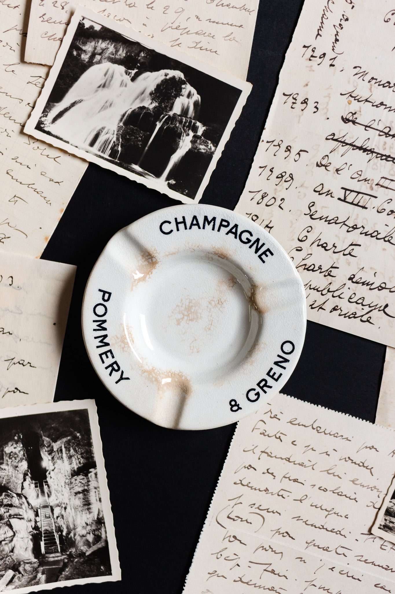 An antique French Luneville Champagne Pommery advertising ashtray rests on handwritten letters and vintage photos, evoking a nostalgic, timeless elegance and a sense of history.