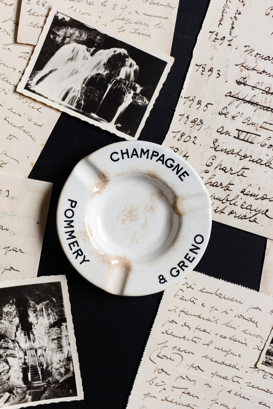An antique French Luneville Champagne Pommery advertising ashtray rests on handwritten letters and vintage photos, evoking a nostalgic, timeless elegance and a sense of history.