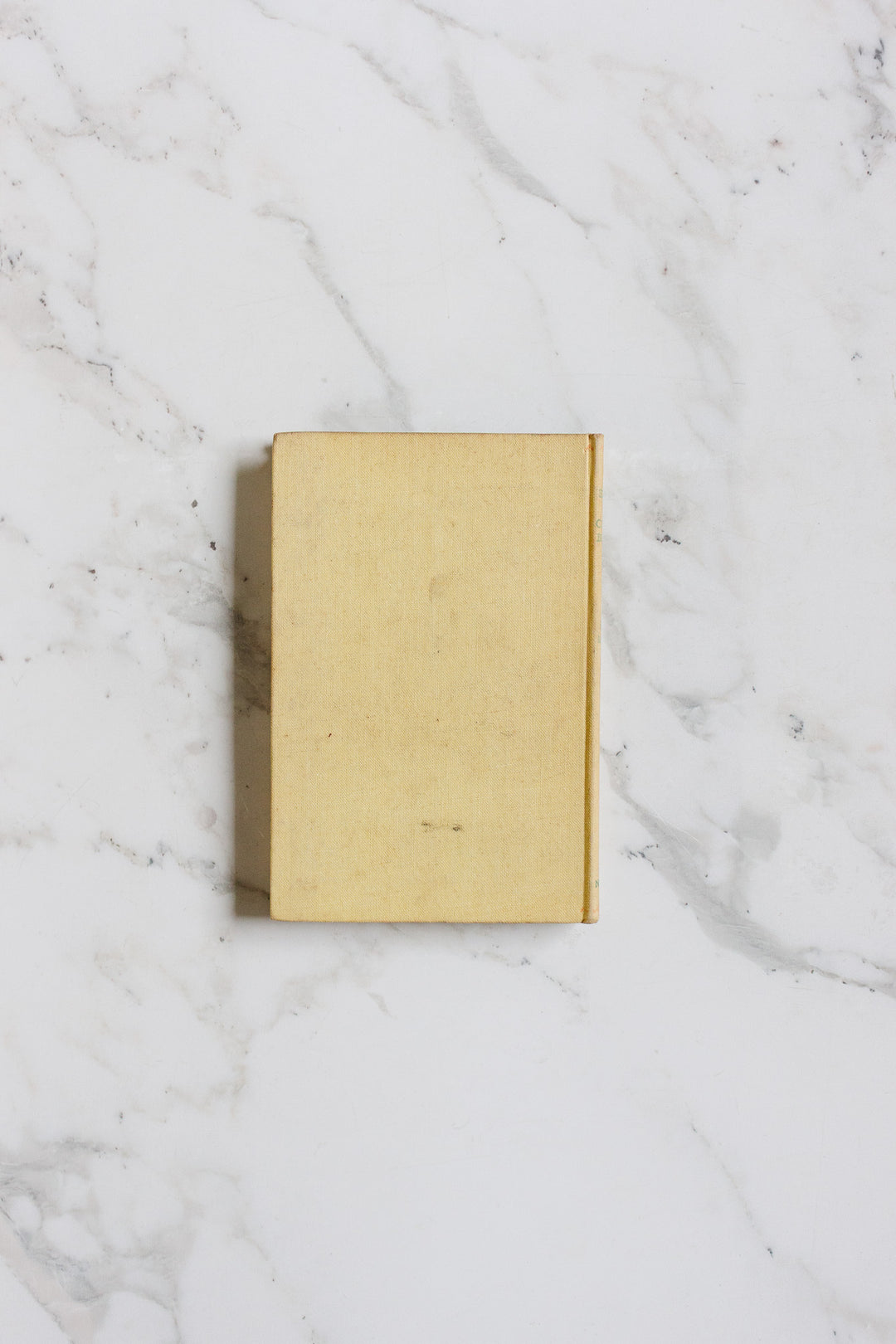 A closed, plain beige hardcover book from books rests on a white marble surface with gray veining. This modest-sized book has no visible title or cover markings.