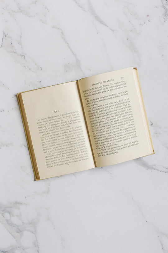 An open book, part of the books collection, rests on a white marble surface with yellowed pages and a beige cover, revealing two pages of French text that reflect its vintage charm and classic appeal.