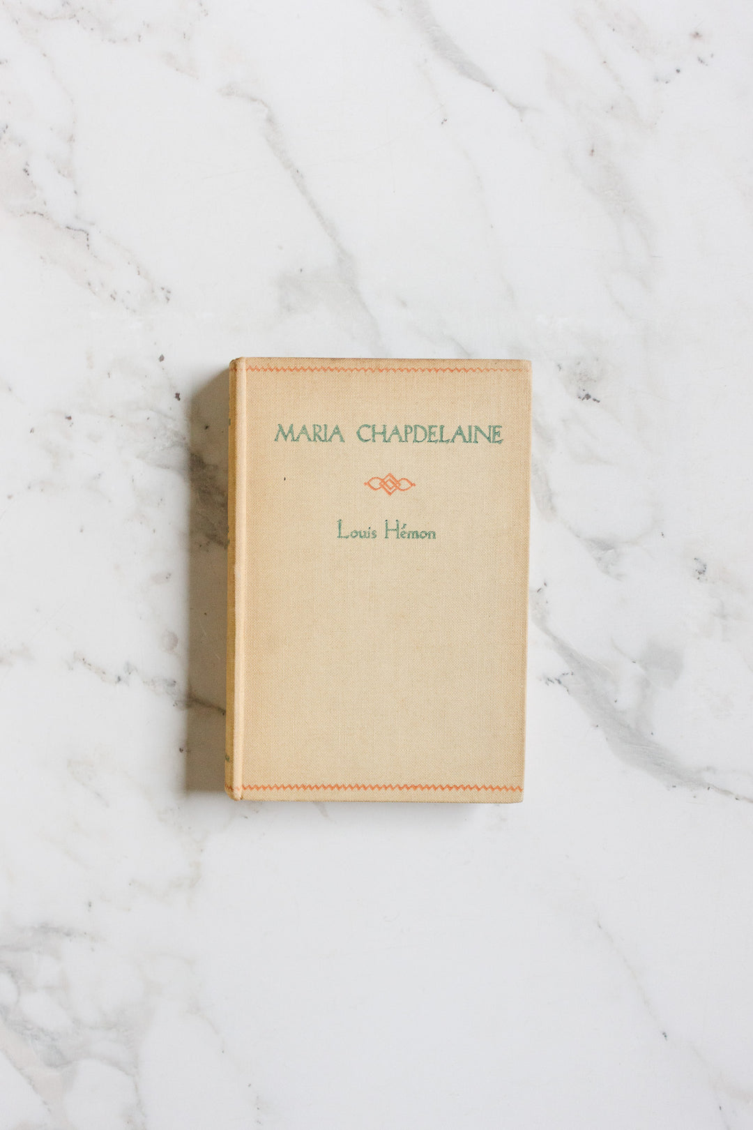A beige hardcover book from the books collection titled "Maria Chapdelaine" by Louis Hémon features the title in green and the author’s name in orange, and sits on a white marble surface—sized to fit any shelf.