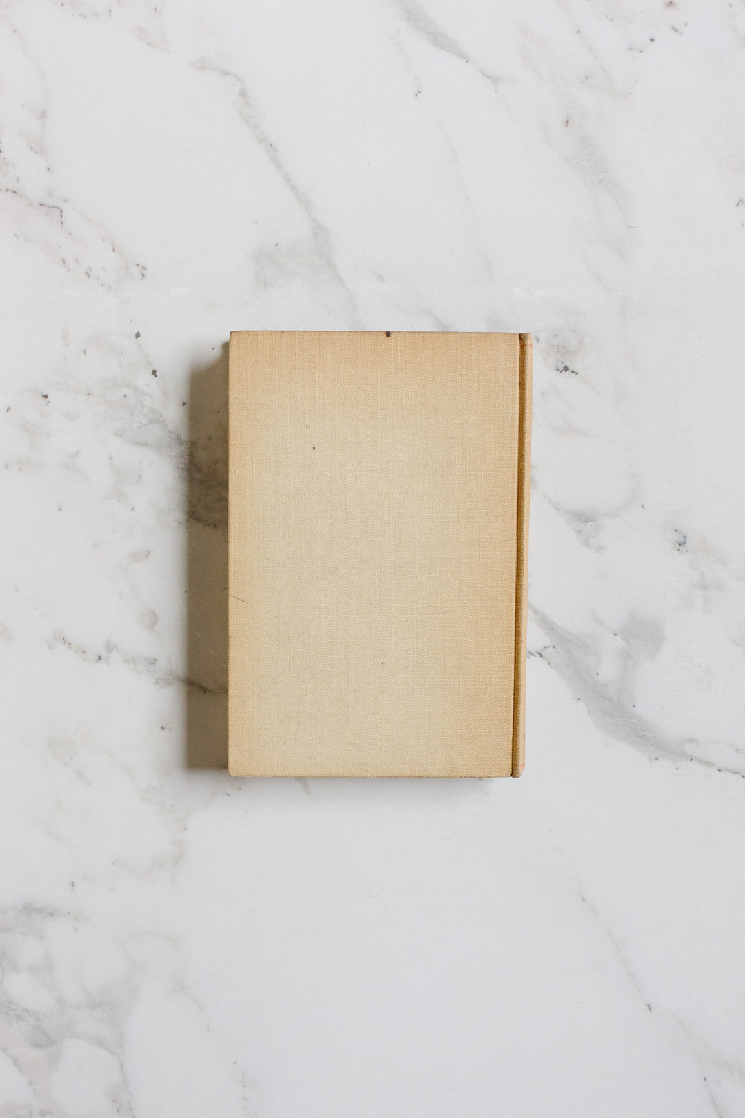 A closed, plain beige hardcover book from Books with no visible title rests on a white marble surface featuring subtle grey veining, highlighted by minimalist composition and clean lines.