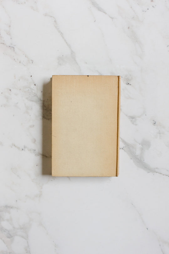 A closed, plain beige hardcover book from Books with no visible title rests on a white marble surface featuring subtle grey veining, highlighted by minimalist composition and clean lines.