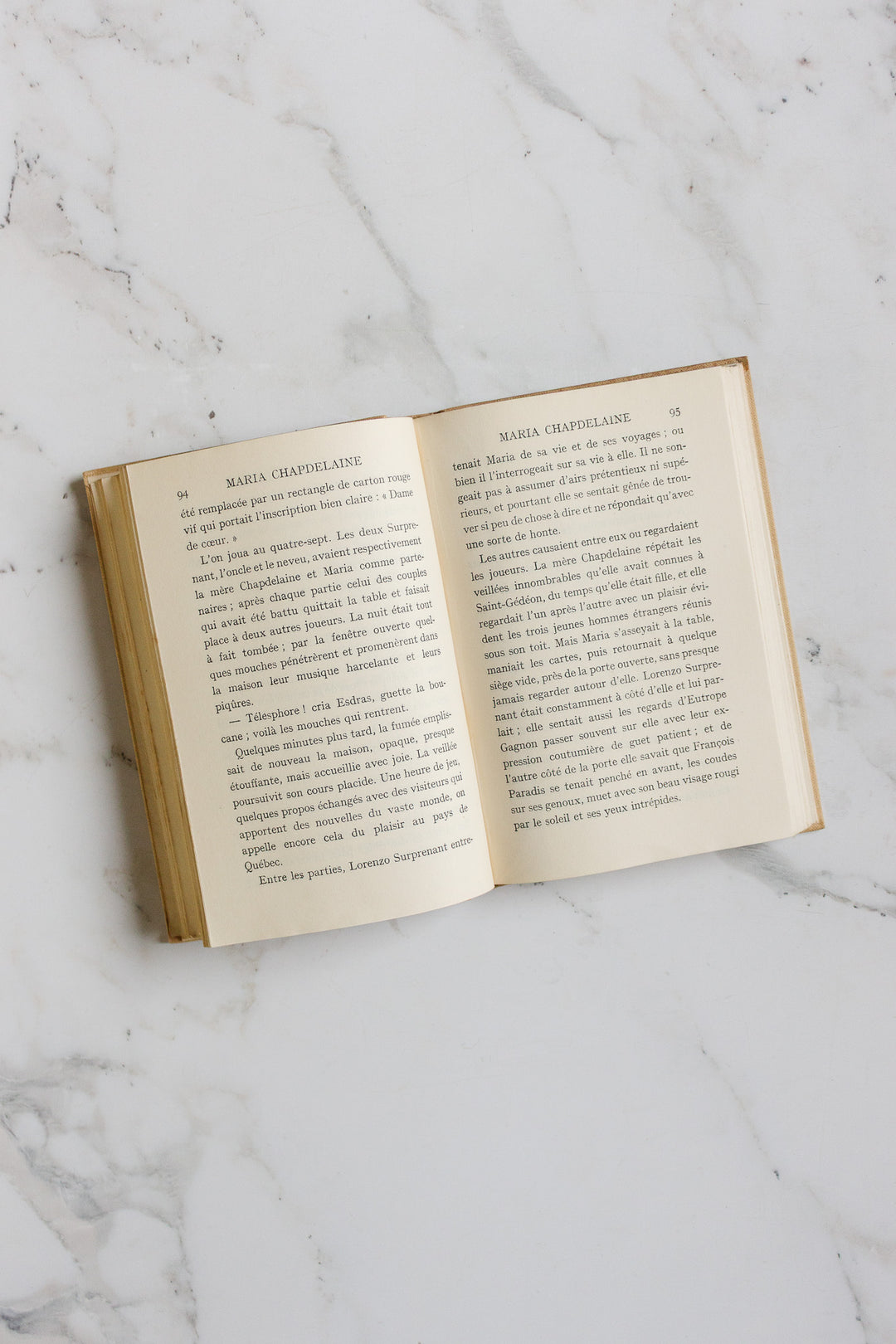 An open hardcover book titled "books" rests on a white marble surface, showing two pages of printed French text; the product name "books" appears at the top of each page.