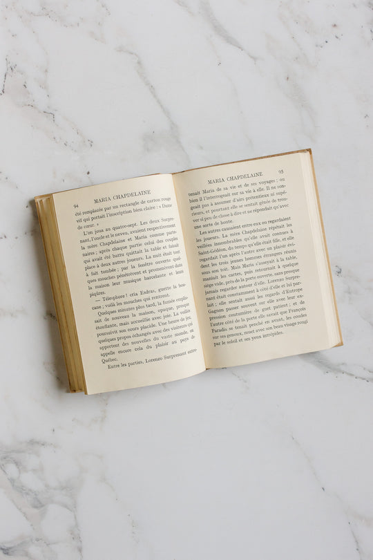 An open hardcover book titled "books" rests on a white marble surface, showing two pages of printed French text; the product name "books" appears at the top of each page.