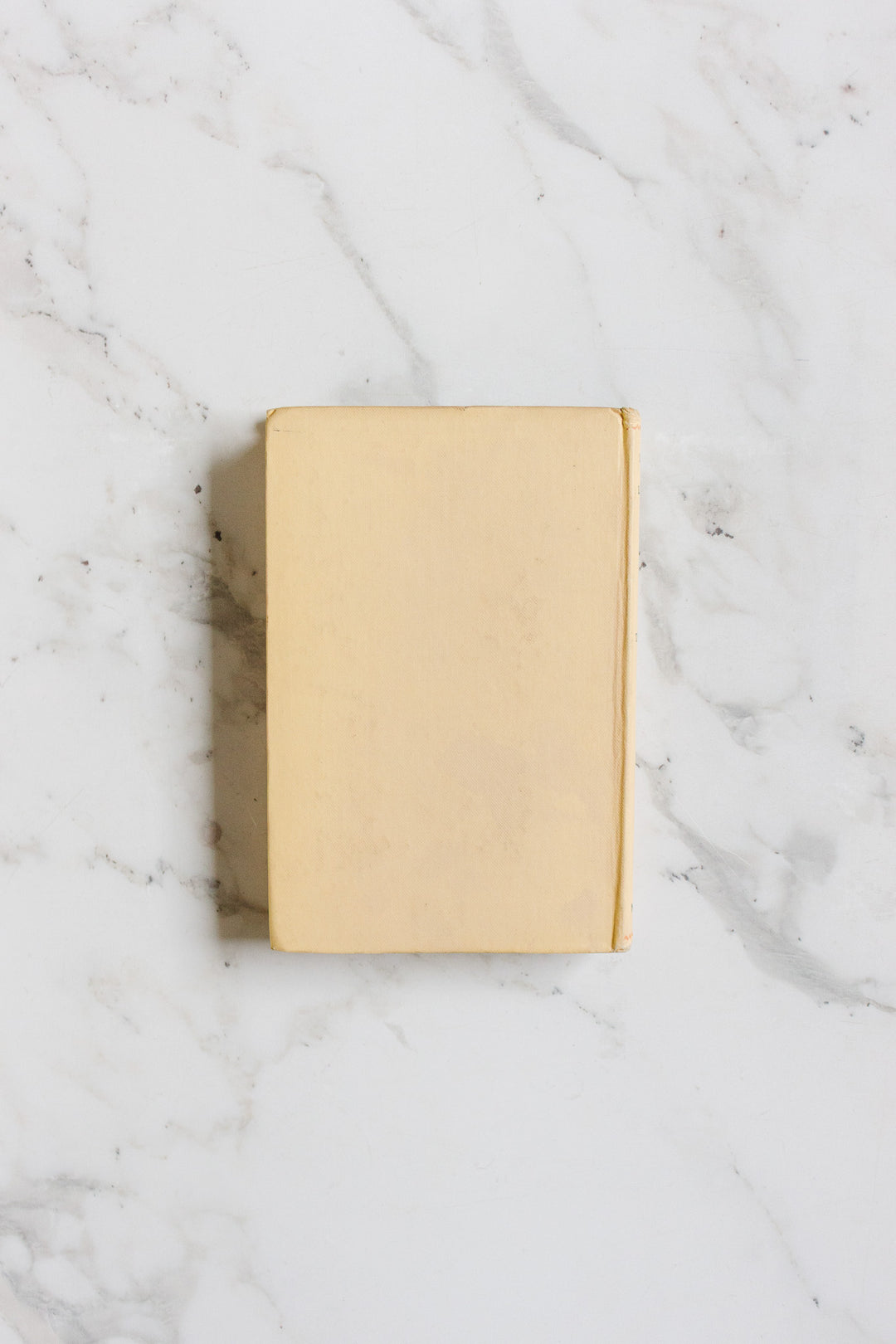A closed, plain beige hardcover book from "books" with discreet dimensions rests on a white marble surface with subtle gray veining.