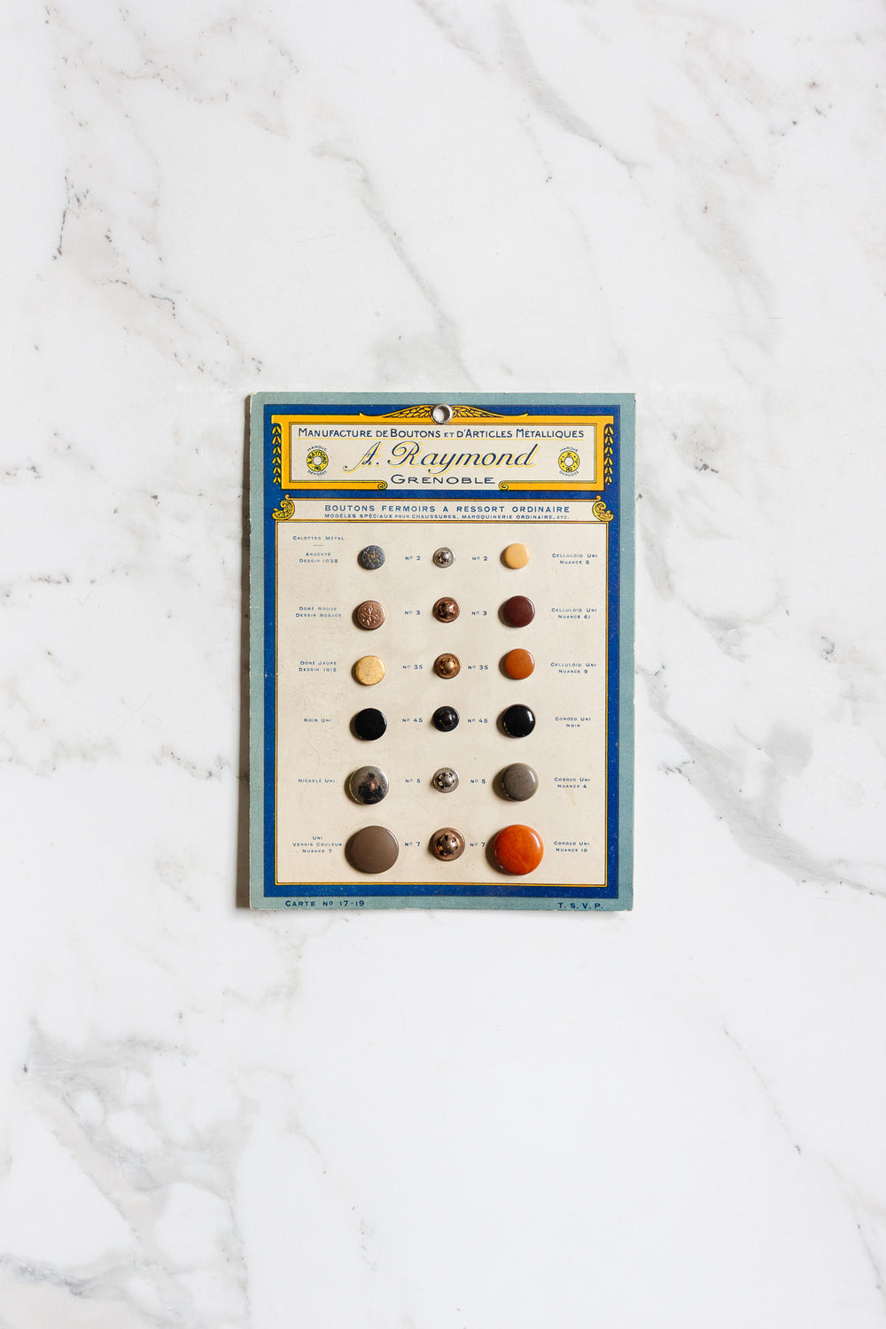A pair of buttons is shown attached to a sample card with French text at the top and a blue and yellow border, displayed on a white marble surface for scale.