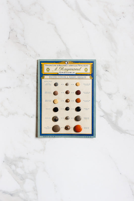 A pair of buttons is shown attached to a sample card with French text at the top and a blue and yellow border, displayed on a white marble surface for scale.