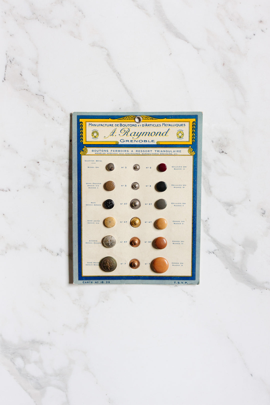 A vintage blue-bordered card labeled "pair of buttons" shows 15 assorted, numbered buttons in various colors and materials on a white marble background, with French text at the top and dimensions for easy reference.