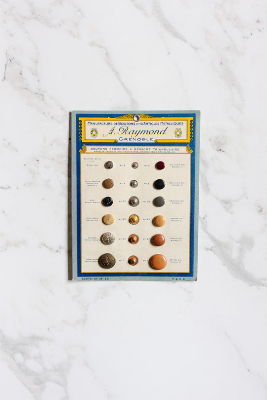 A vintage blue-bordered card labeled "pair of buttons" shows 15 assorted, numbered buttons in various colors and materials on a white marble background, with French text at the top and dimensions for easy reference.