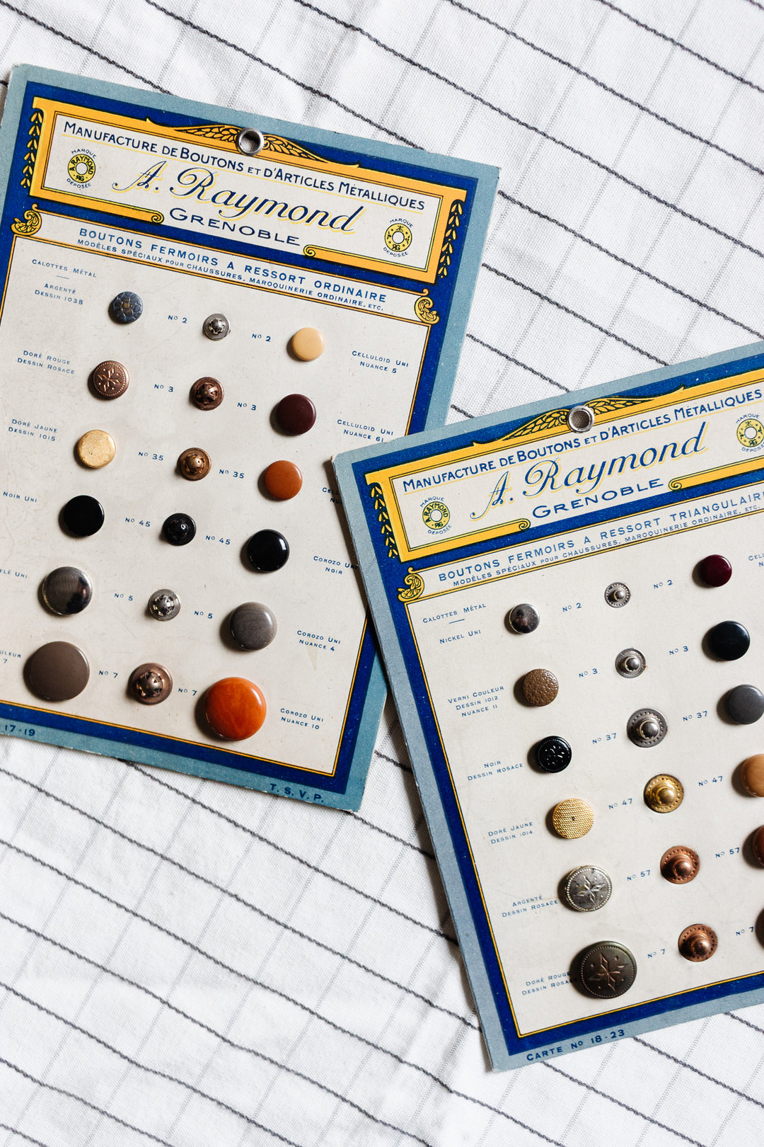 A pair of buttons is displayed on vintage sample cards labeled J. Raymond Grenoble, each metal button attached and neatly arranged on a white cloth with a grid pattern for scale.