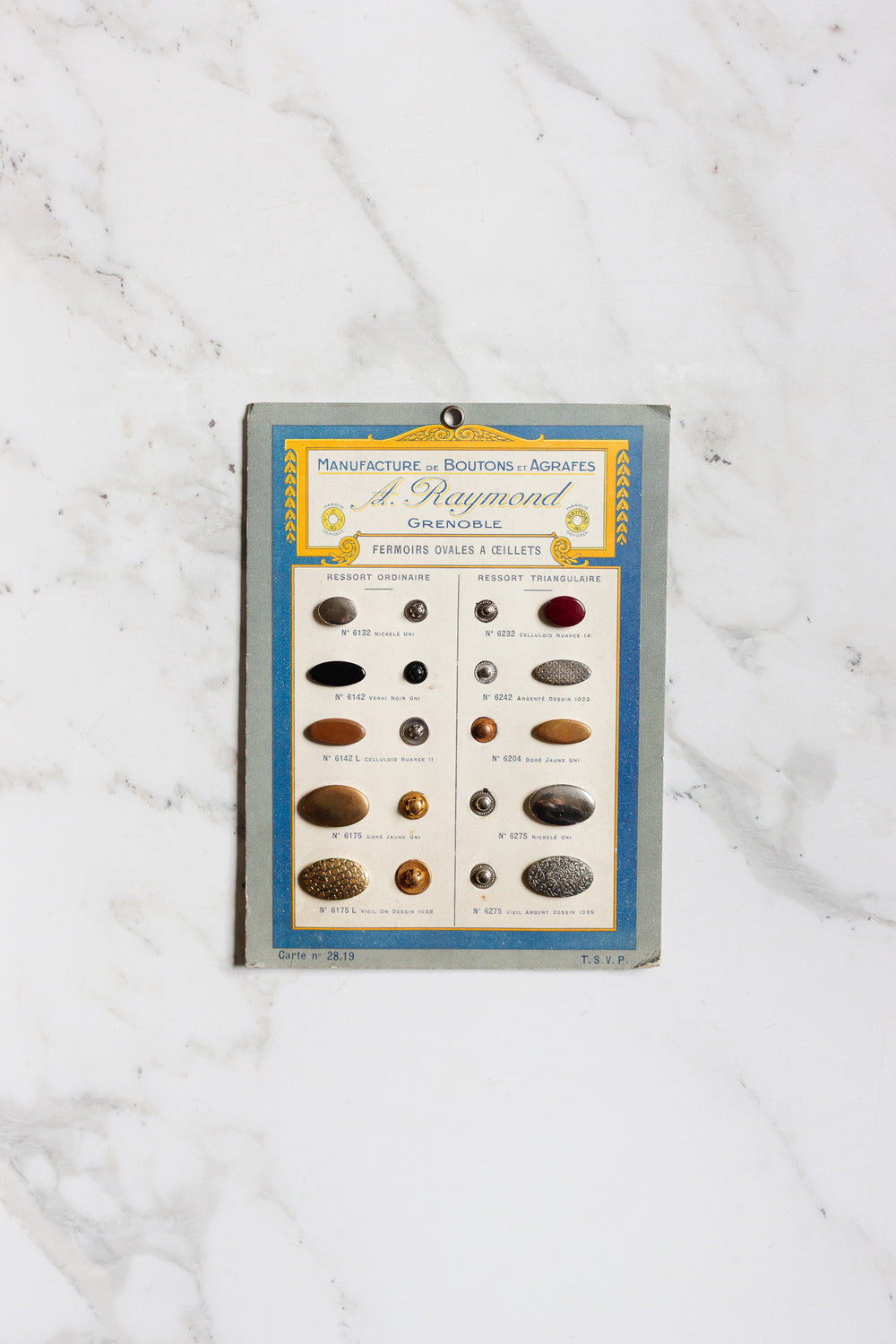 A 1920s A. Raymond sample card from Grenoble displays assorted metal and fabric buttons with labeled descriptions, including vintage button clasps, set on a white marble background.
