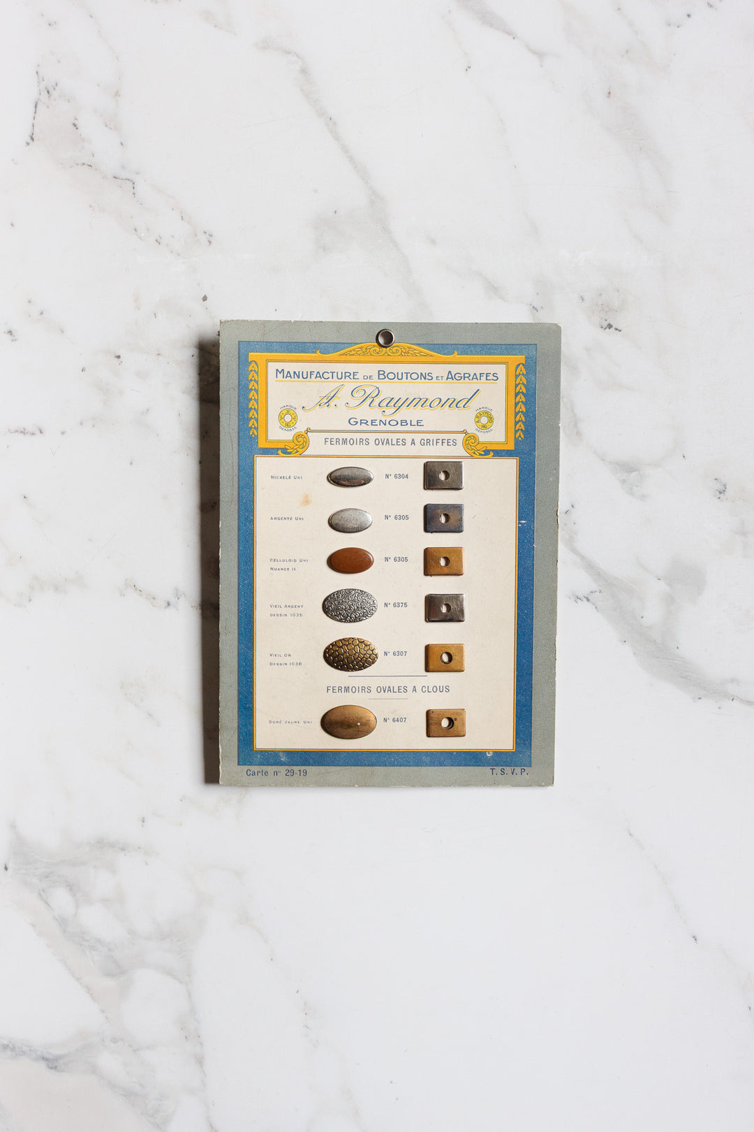 Pair of buttons ii: A vintage card featuring assorted oval and round metal buttons and fasteners labeled in French, beautifully set on a classic white marble background.
