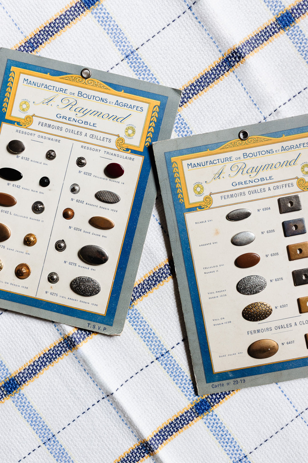 Two 1920s sample cards from A. Raymond, manufacture de boutons et agrafes - Grenoble, display vintage button clasps in assorted styles and colors on a white, blue, and yellow checkered fabric background.