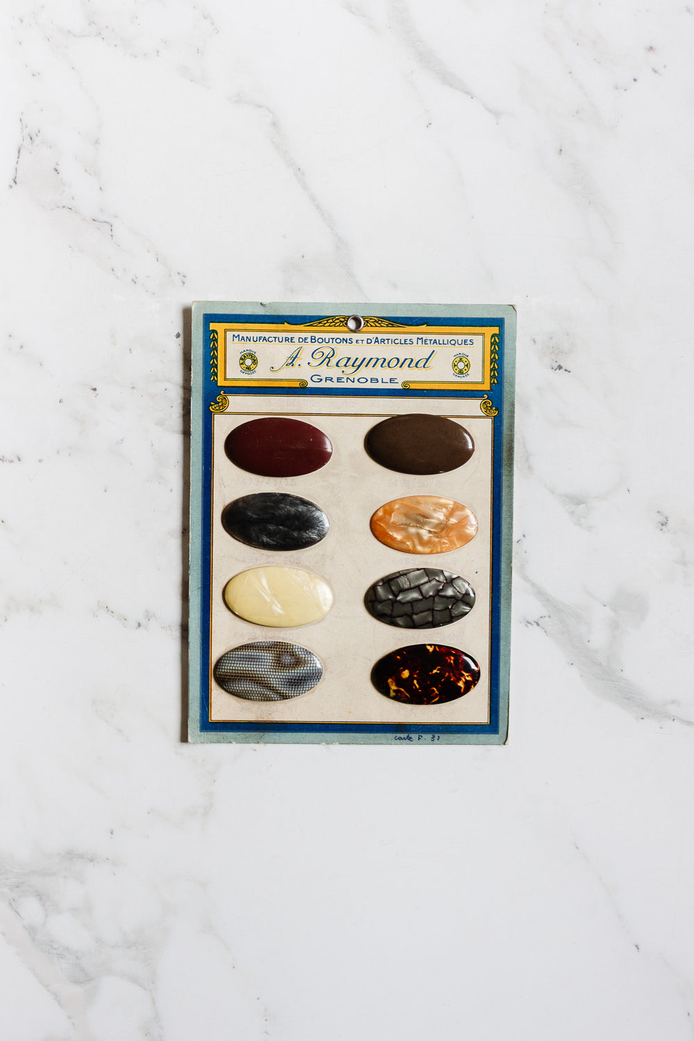 A vintage card displays seven assorted 1920s antique A. Raymond buttons – Grenoble, in various colors and patterns, arranged in two vertical columns on a white marble surface. The card has a blue border and French text at the top.