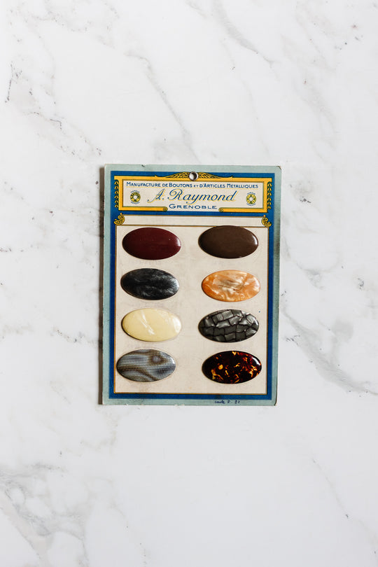 A vintage card displays seven assorted 1920s antique A. Raymond buttons – Grenoble, in various colors and patterns, arranged in two vertical columns on a white marble surface. The card has a blue border and French text at the top.