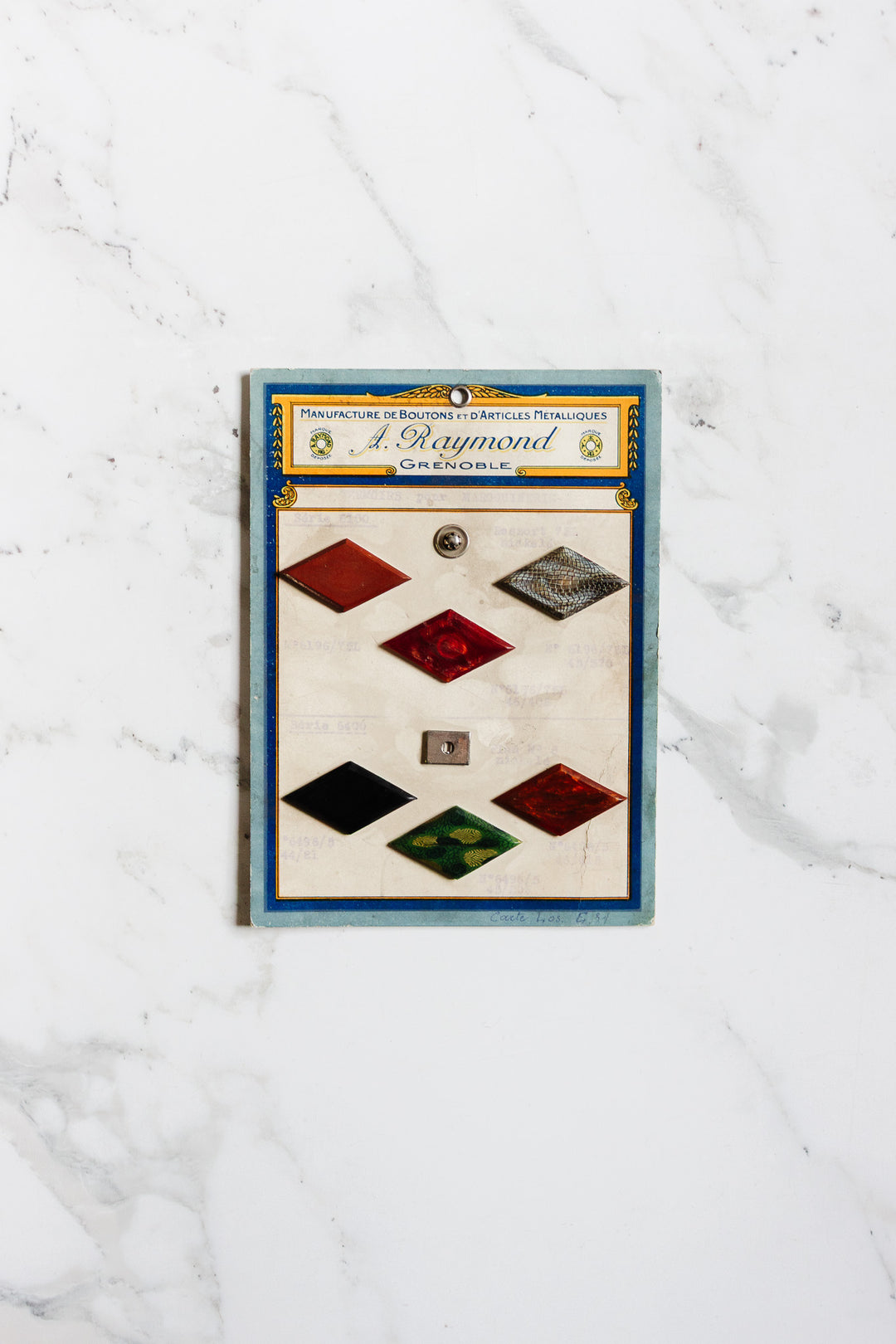A vintage card of 1920s antique A. Raymond buttons – grenoble, featuring seven assorted buttons in different shapes, colors, and sizes on a white marble background. The card has a blue border and French text inspired by early 20th-century French fashion.