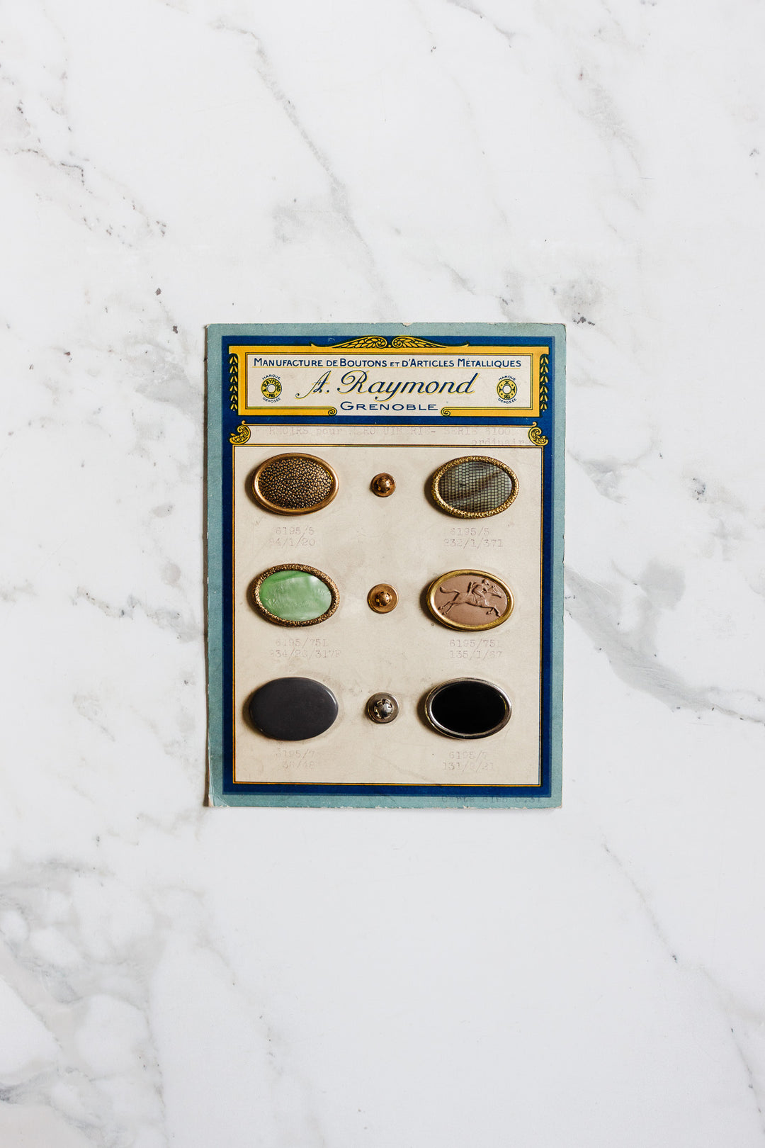 A vintage card featuring six unique 1920s antique A. Raymond buttons from Grenoble, each with assorted designs and colors, elegantly displayed on a white marble surface—an ode to early 20th-century French style.