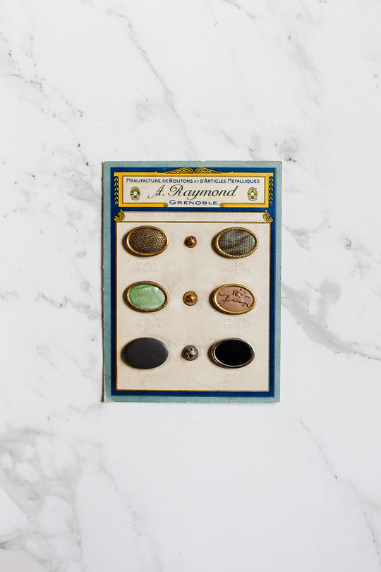 A vintage card featuring six unique 1920s antique A. Raymond buttons from Grenoble, each with assorted designs and colors, elegantly displayed on a white marble surface—an ode to early 20th-century French style.
