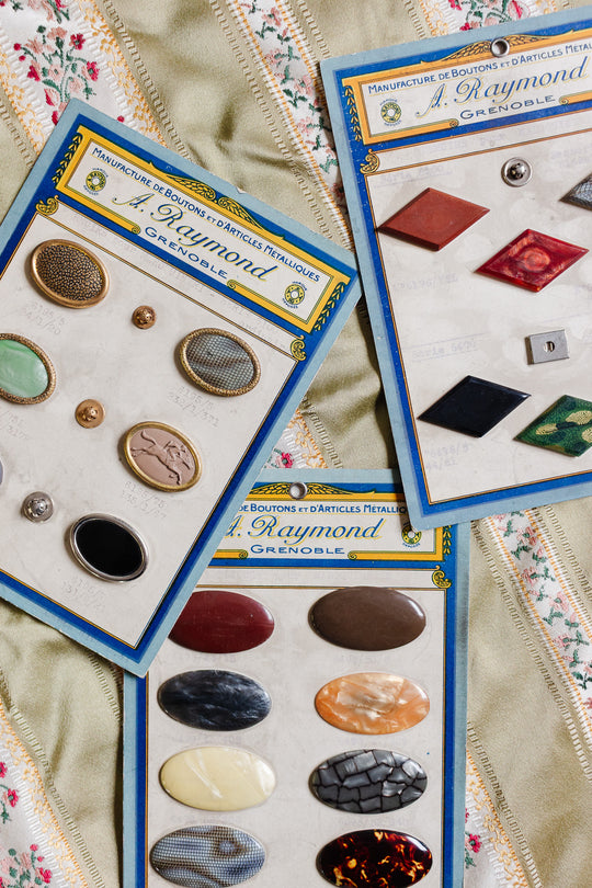 Three 1920s antique A. Raymond – Grenoble button cards display assorted French vintage buttons in various shapes, colors, and materials, highlighting early 20th-century French fashion on a patterned fabric background.
