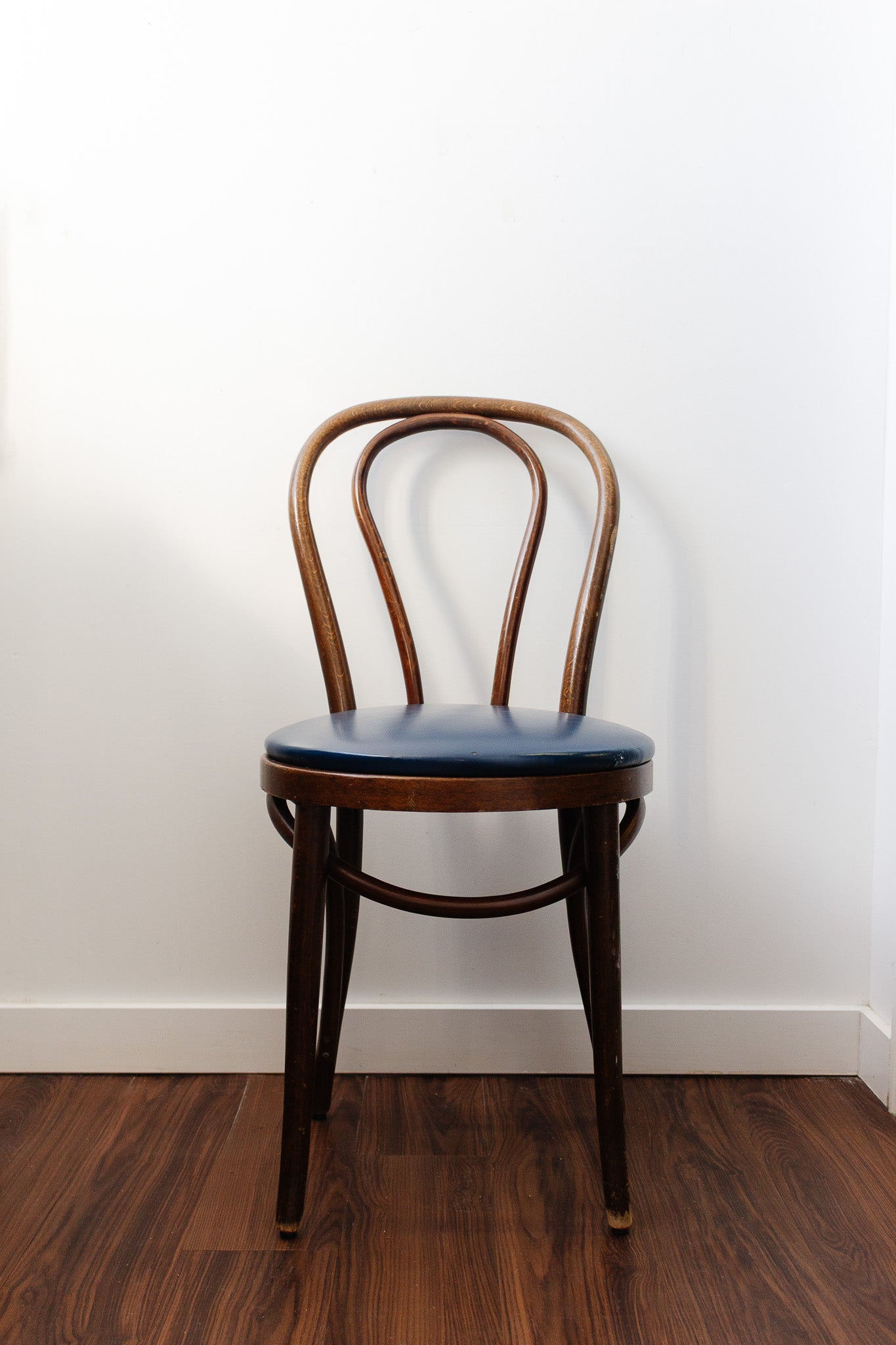 A vintage pair of Thonet bentwood bistro chairs with curved backrests and dark blue seat cushions sits on a wooden floor against a plain white wall.