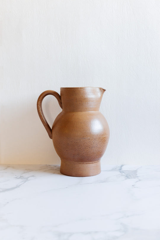 The large jug, a brown ceramic pitcher with a handle and classic proportions, rests on a white marble surface against a light textured background.