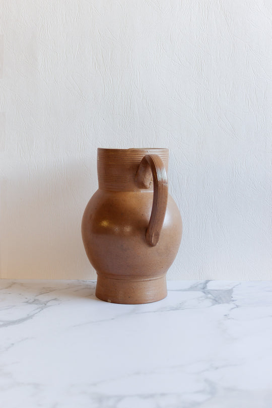 The large jug, crafted from brown ceramic with a single handle and classic proportions, rests on a white marble surface against a textured white backdrop.