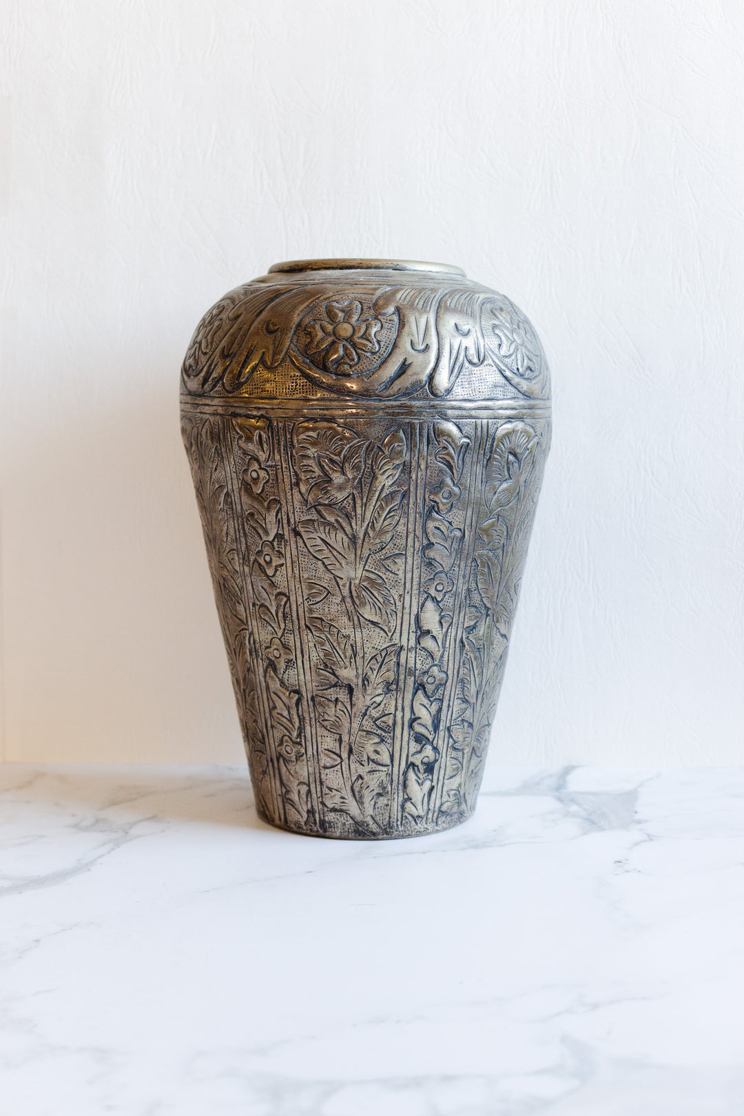 A tall, ornate metal vase with detailed floral and leaf engravings stands elegantly on a white marble surface against a simple, light background.