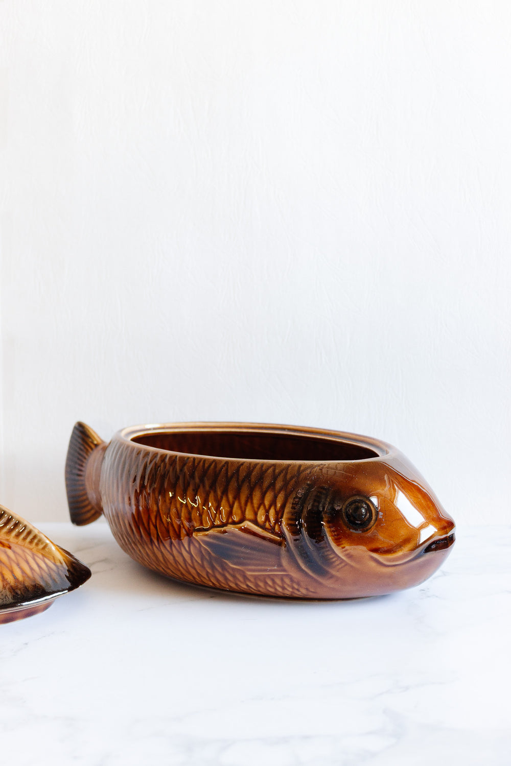 The fish soupiere is a ceramic serving dish with a glossy brown finish, shaped like a fish. It comes with a matching lid and is displayed on a white surface. Check the dimensions to ensure it fits your table setting perfectly.