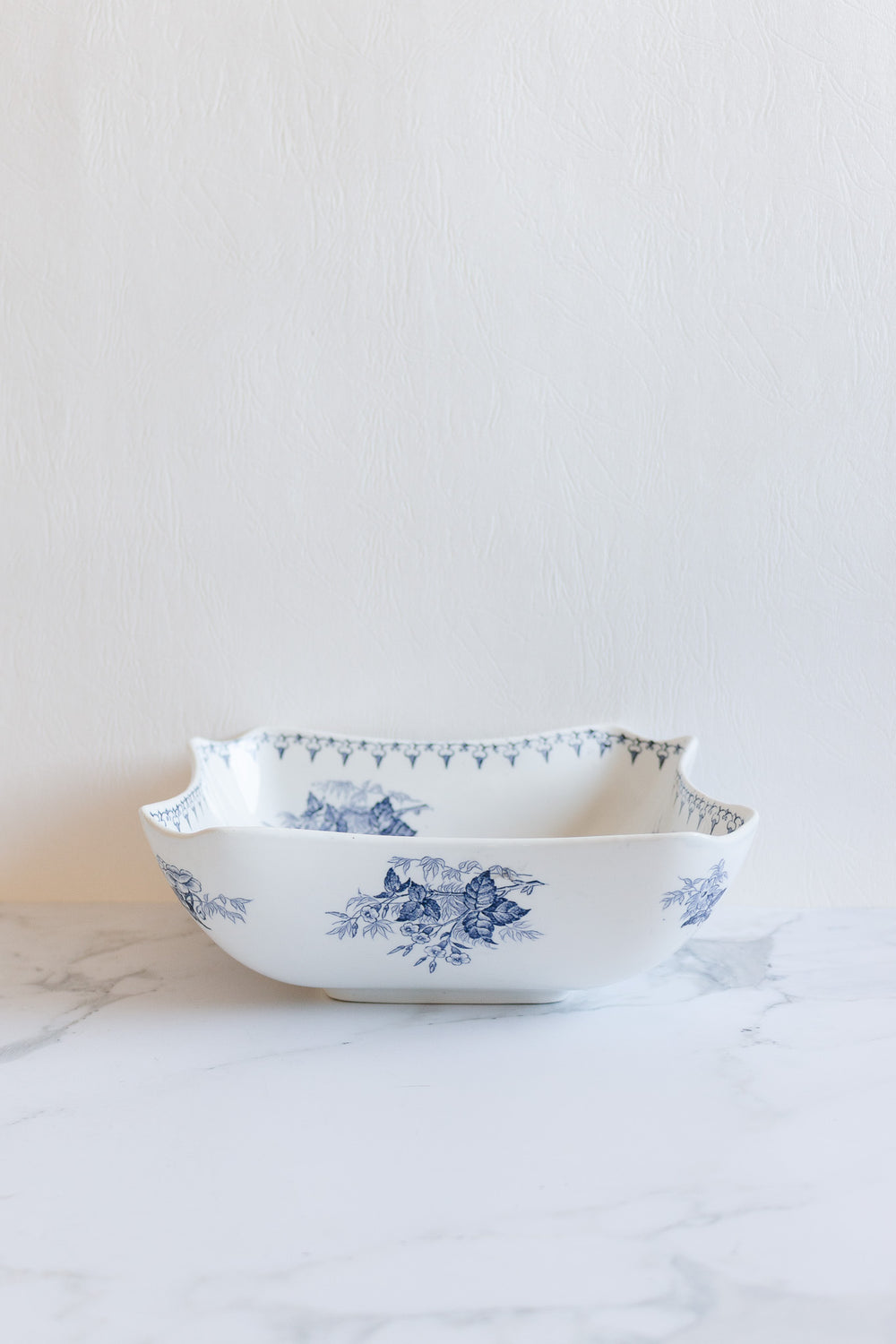 The floral bowl features blue floral patterns on white ceramic along the sides and rim, shown on a marble surface with a light textured backdrop. Contact us for exact dimensions.