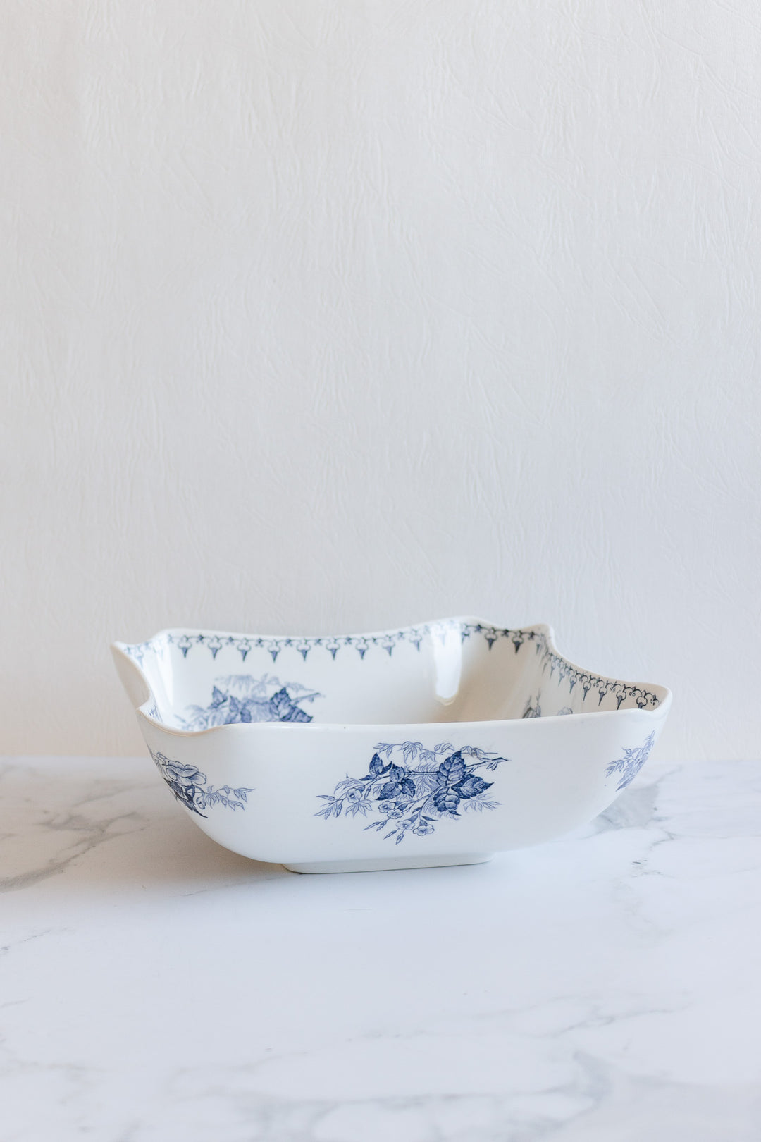 The floral bowl is a white ceramic dish with blue floral patterns and gently scalloped edges. Elegant yet practical, it’s ideal for display or daily use, shown here on a white marble surface against a light textured background.