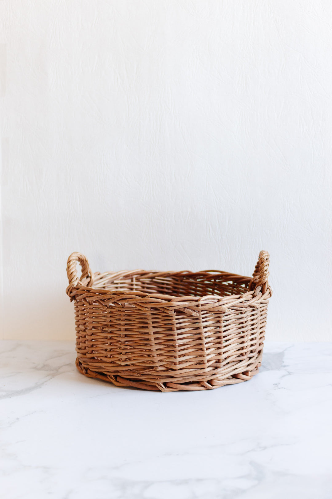 The basket, a spacious round wicker piece with two side handles, sits empty on a white marble surface against a light textured background.