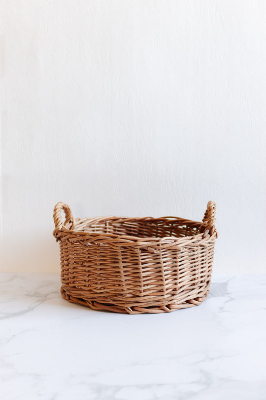 The basket, a spacious round wicker piece with two side handles, sits empty on a white marble surface against a light textured background.