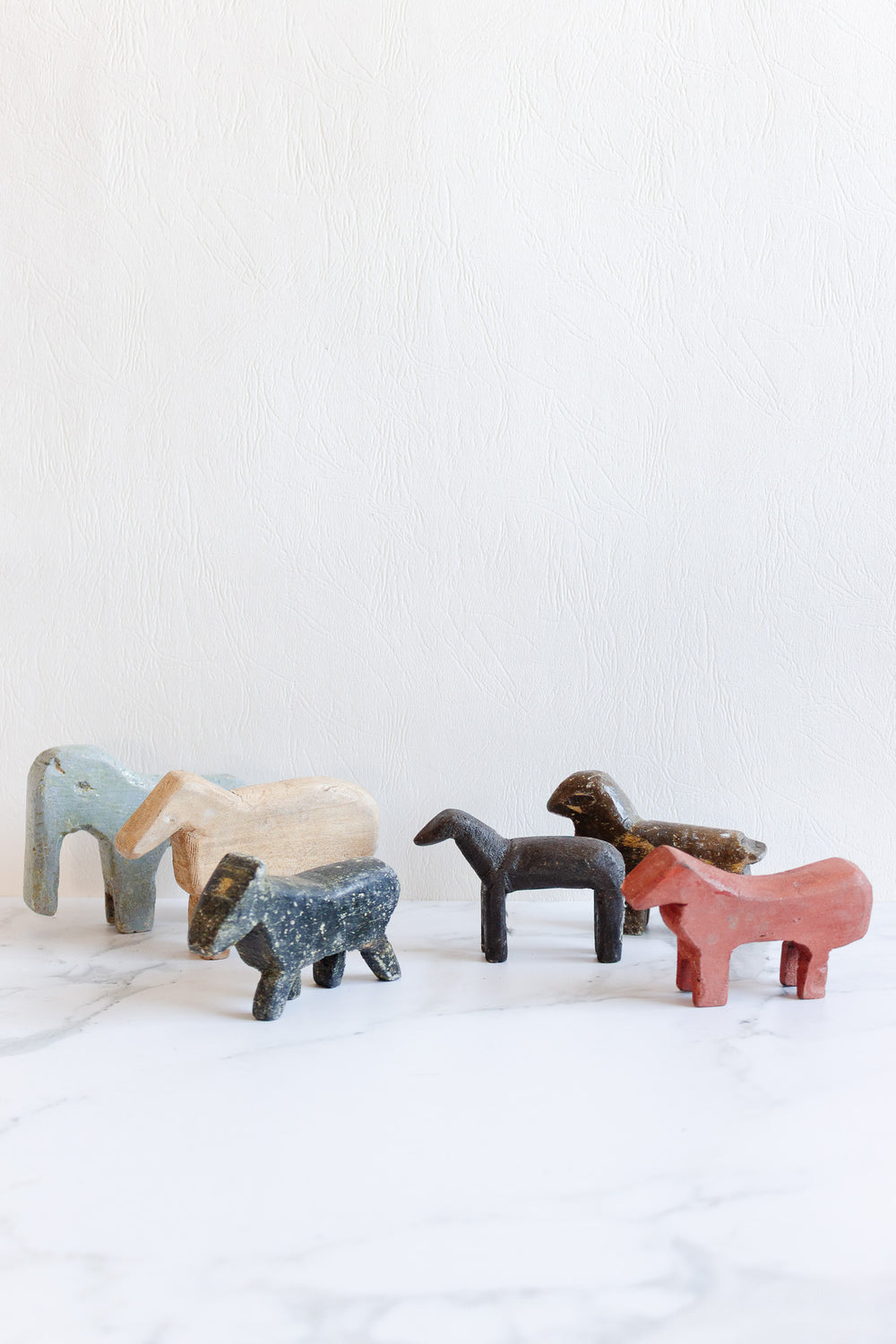 Six colorful, minimalist wooden animals—each uniquely sized and shaped—stand on a white marble surface against a textured white background. The figures resemble horses or donkeys in different poses.