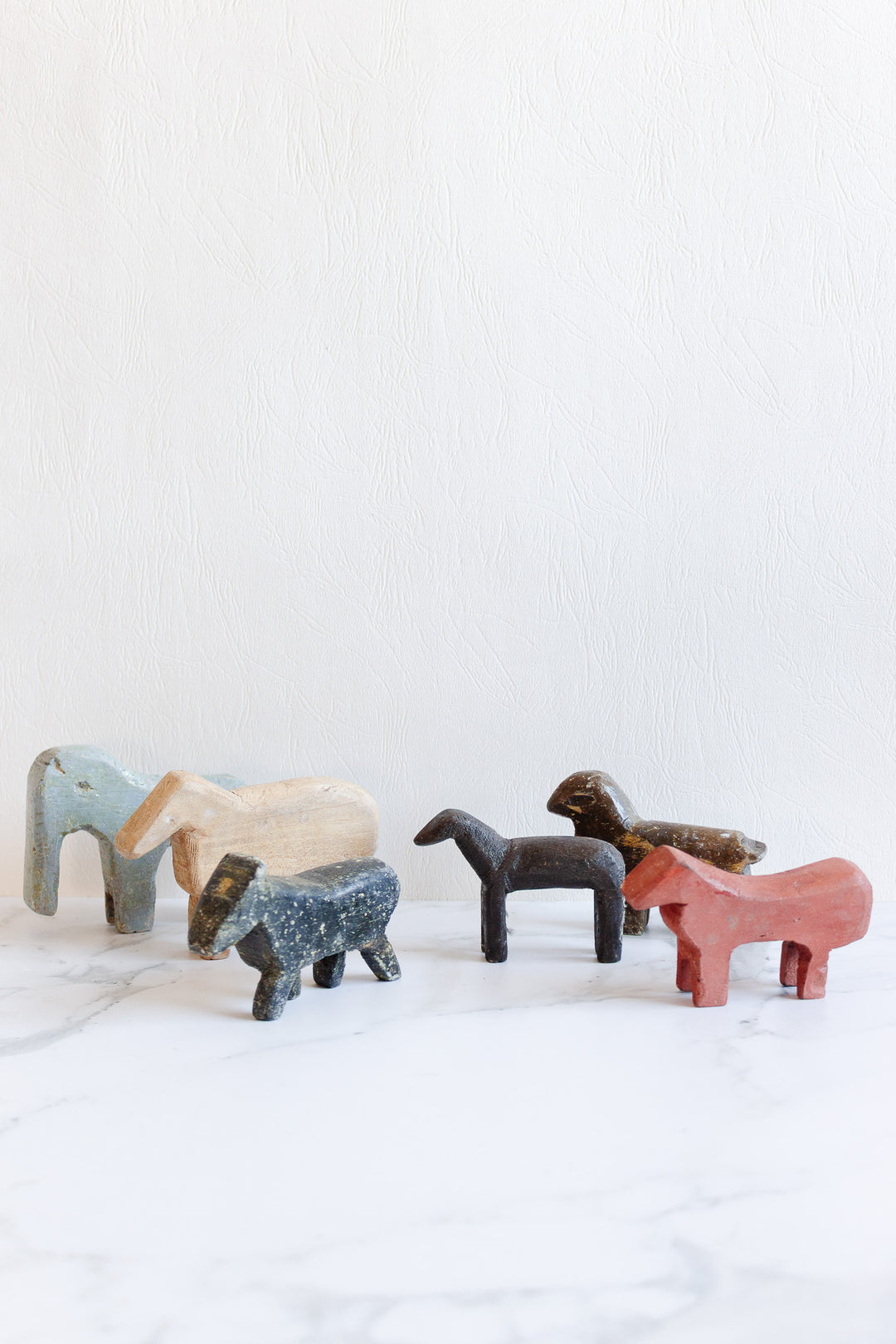Six colorful, minimalist wooden animals—each uniquely sized and shaped—stand on a white marble surface against a textured white background. The figures resemble horses or donkeys in different poses.