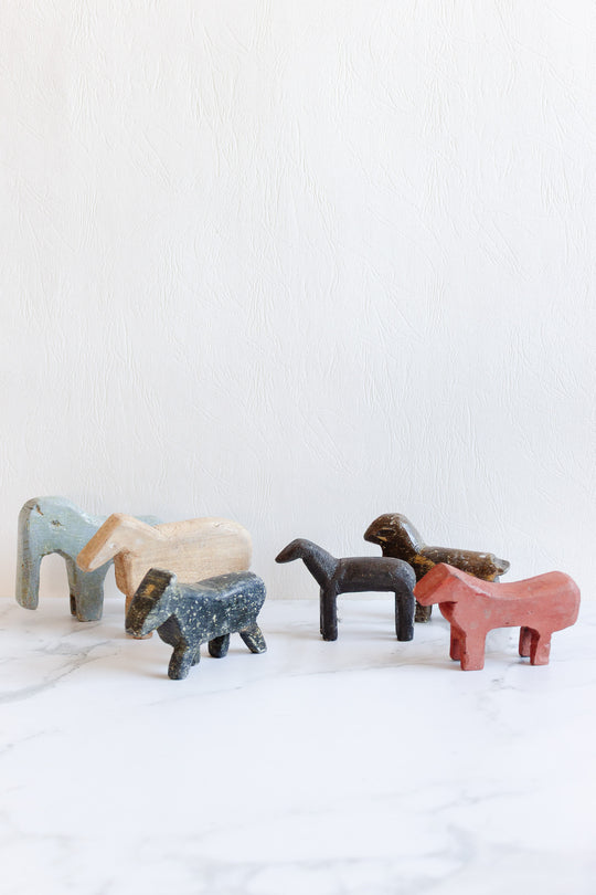 Six colorful, minimalist wooden animals—each uniquely sized and shaped—stand on a white marble surface against a textured white background. The figures resemble horses or donkeys in different poses.