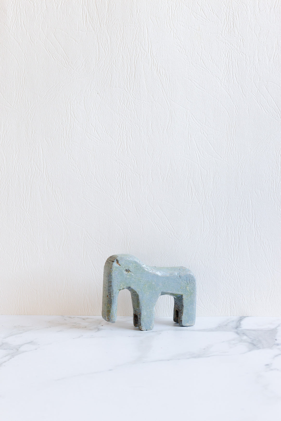 A compact, abstract blue-gray wooden animal sculpture stands on a white marble surface against a textured white wall, its minimalist design enhancing its charm.