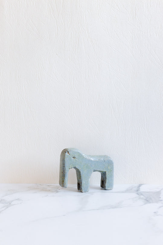 A compact, abstract blue-gray wooden animal sculpture stands on a white marble surface against a textured white wall, its minimalist design enhancing its charm.
