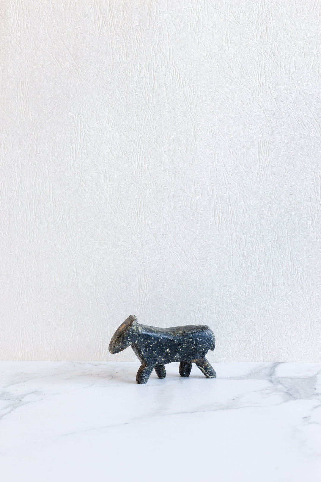 A small wooden animals figurine with four legs and a circular head rests on a white marble surface against a textured white wall, its compact size ideal for tabletop display.