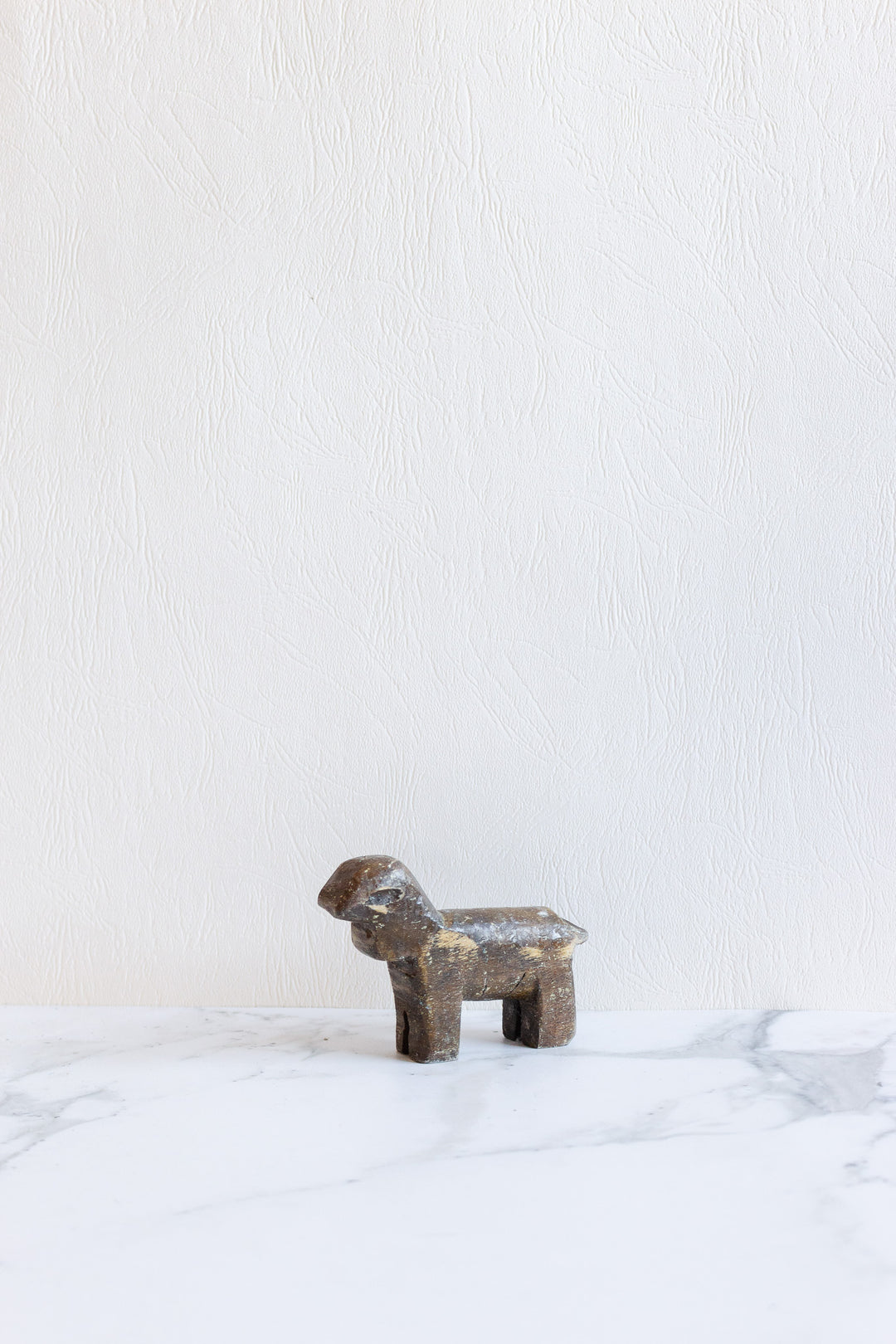A small, rustic dark brown wooden animals figurine sits on a white marble surface, its form standing out against a textured off-white wall.