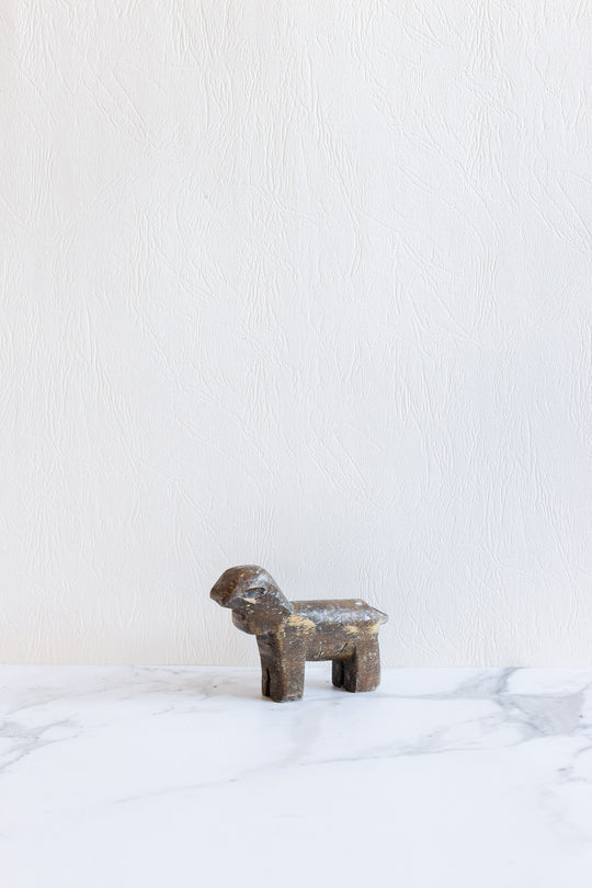 A small, rustic dark brown wooden animals figurine sits on a white marble surface, its form standing out against a textured off-white wall.