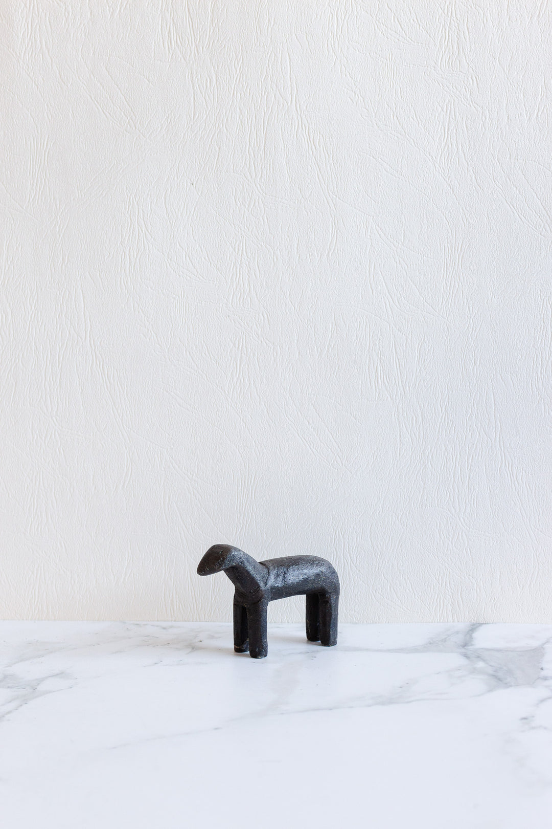 A compact, minimalist black wooden animal sculpture with four legs stands on a white marble surface against a textured white wall—its small size is perfect for modern decor.