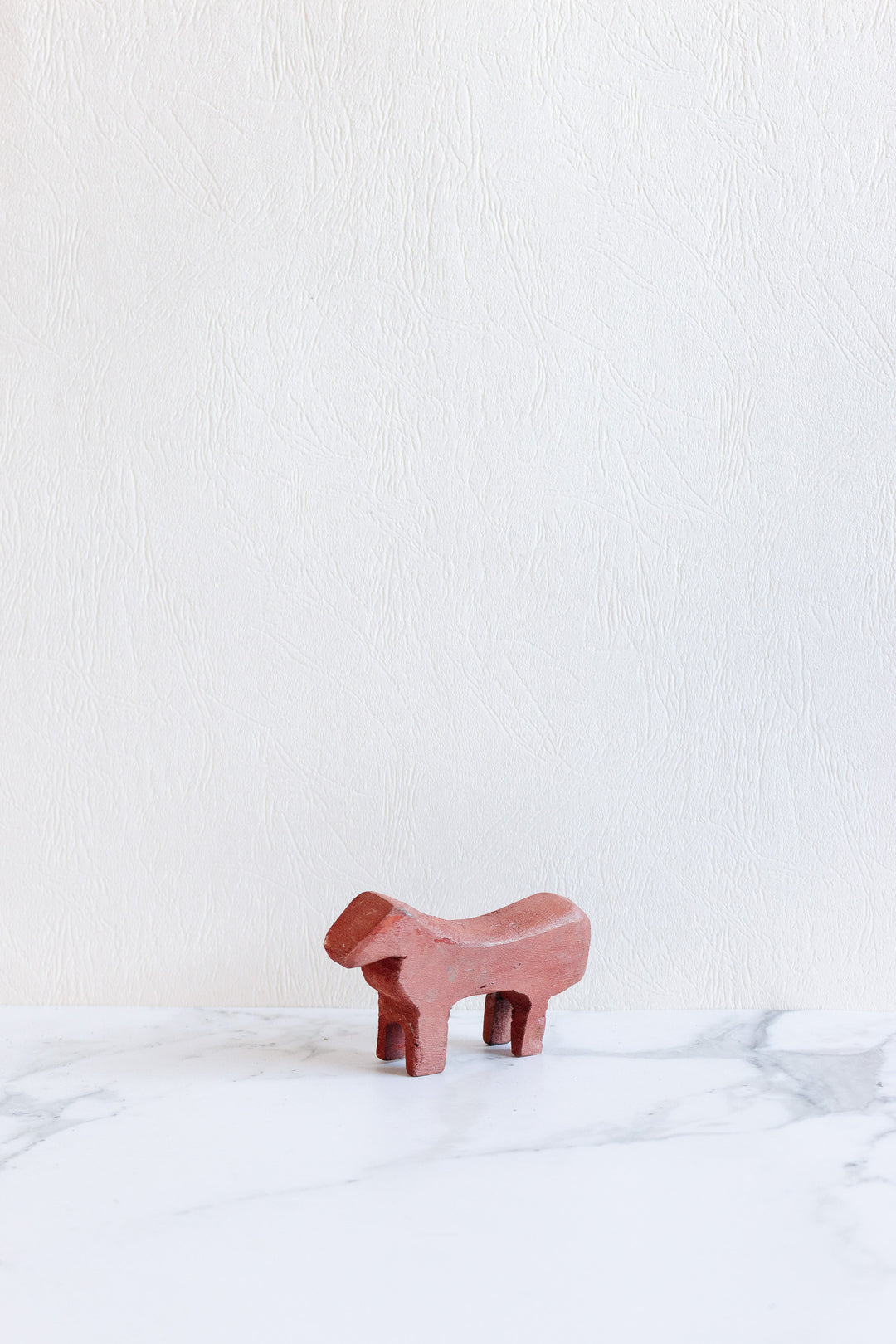 A small, minimalist wooden animals figurine in reddish-brown stands on a white marble surface against a textured light background.