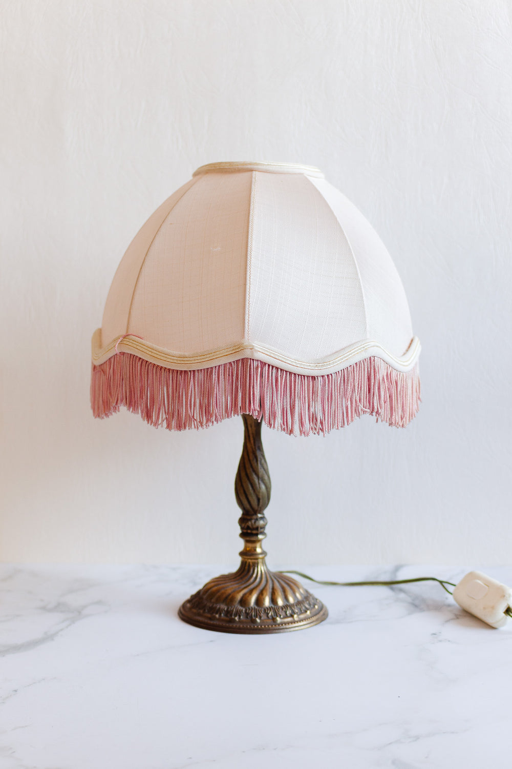 A vintage lamp with a brass base and a cream fabric shade accented by pink fringe rests on a white marble surface. Its dimensions and electric cord with switch are also visible beside the lamp.