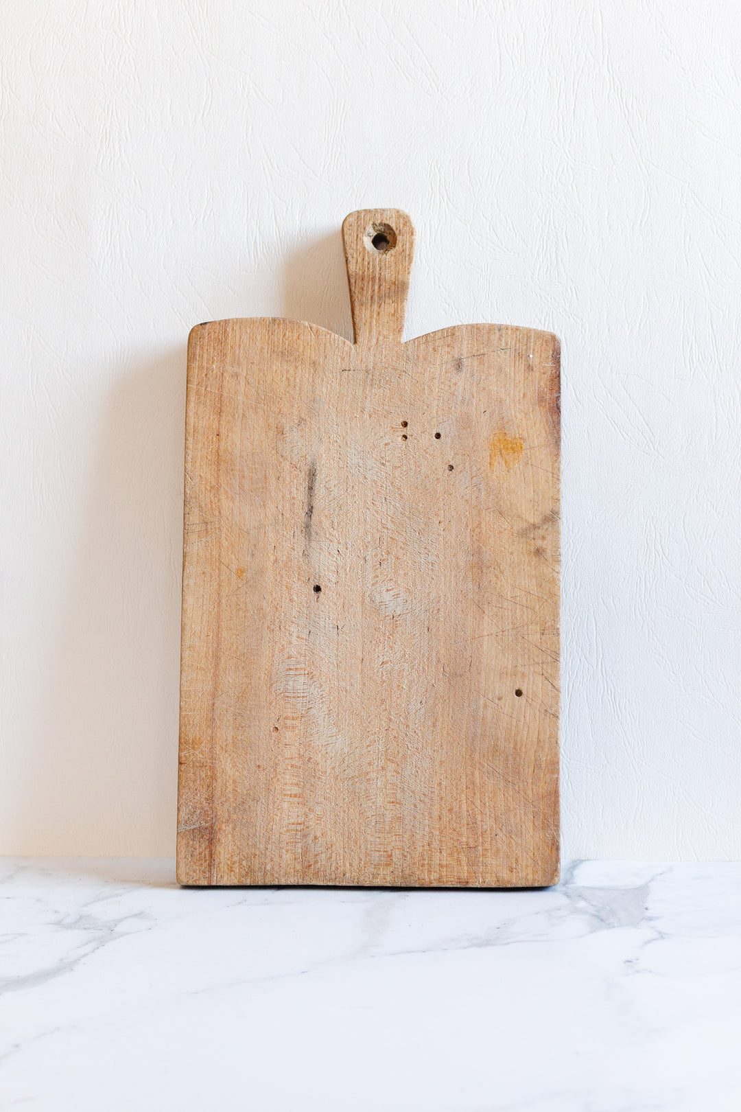 A breadboard with a handle and hole, featuring sleek rectangular dimensions, stands upright against a white textured wall on a white marble surface.