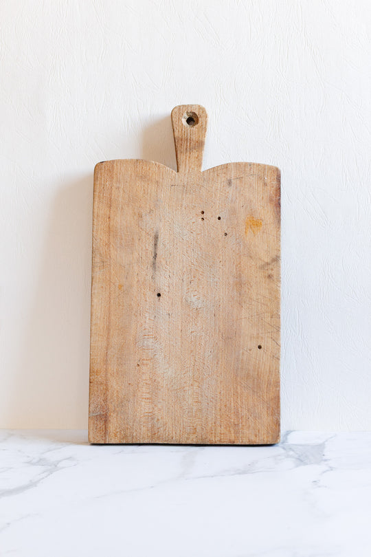 A breadboard with a handle and hole, featuring sleek rectangular dimensions, stands upright against a white textured wall on a white marble surface.