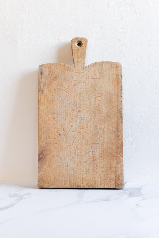 A breadboard with a handle and hanging hole stands upright against a white textured wall on a marble surface. Its practical rectangular design is ideal for everyday kitchen tasks.